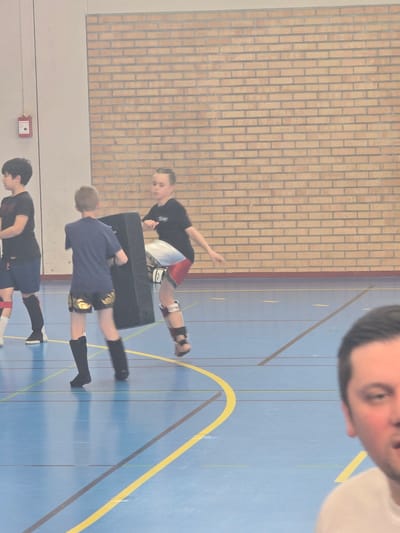 Three children, wearing athletic apparel and shin guards, are practicing kicks in a gymnasium with a blue floor marked with yellow and brown lines. A black padded target sits in front of the top child. The gymnasium walls are a mix of brick and painted surfaces. The location is likely Leers, France. A man's face appears in the upper right corner of the image.
