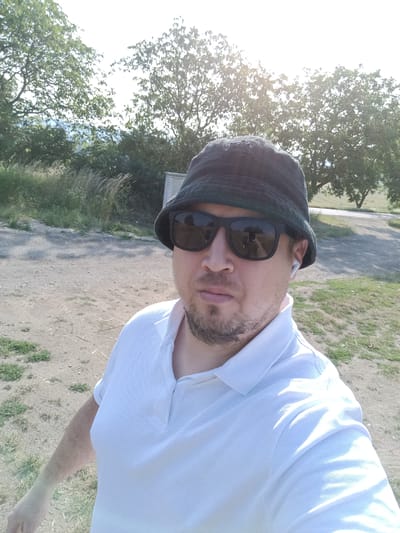 This is a selfie taken by a man outdoors on a sunny day. He is wearing a dark bucket hat, sunglasses, and a white polo shirt. He also has white wireless earbuds in his ears. The man has a light beard and appears to be in his 30s or 40s.
The background shows trees and dry grass, suggesting a rural or park-like setting. The ground is covered in dirt and gravel, with some patches of green grass. The sun is bright, casting shadows and creating a slight glare in the image, indicating it is likely daytime, possibly late morning or early afternoon.
The overall mood of the image is casual and relaxed. The man is looking directly at the camera, and his expression is neutral. There is no visible text in the image. The location context provided is Šternov, Czechia.