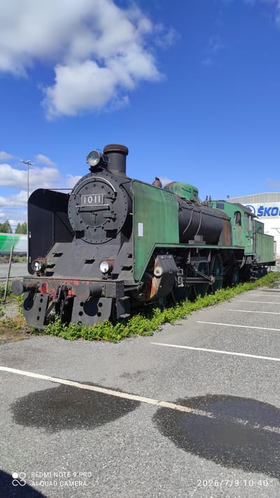 A weathered, green and black steam locomotive, numbered "1011", is parked in a paved lot outside under a bright blue sky with scattered white clouds. The locomotive, which appears to be a static display, is surrounded by green vegetation at its base. In the background, a white building with a blue "ŠKODA" logo is visible. The ground in the foreground is asphalt, marked with white lines indicating parking spaces, and several dark, wet patches, likely puddles. The scene is captured in daylight.