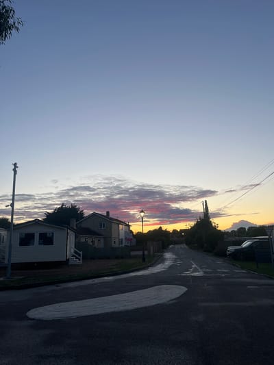 This image depicts a street scene in Harwich, United Kingdom, during twilight. The sky displays a gradient of blues and purples with streaks of pink and orange from the setting sun. A few clouds are visible in the distance, illuminated by the sunset.

On the left side of the road, there are two structures. The closest one is a white mobile home or caravan, with windows displaying "I'm For Sale" signs. Next to it is a more traditional house, a two-story building with a pitched roof. A street lamp stands on the pavement between these two structures.

The road itself appears wet, with reflections from the sky and street lighting. There is a speed bump painted in a light color on the road. Further down the street, trees and bushes line the sides, and power lines are visible overhead.

On the right side of the road, two dark-colored cars are parked. A "Give Way" traffic sign is also visible. The overall impression is a quiet residential street as day transitions into night.