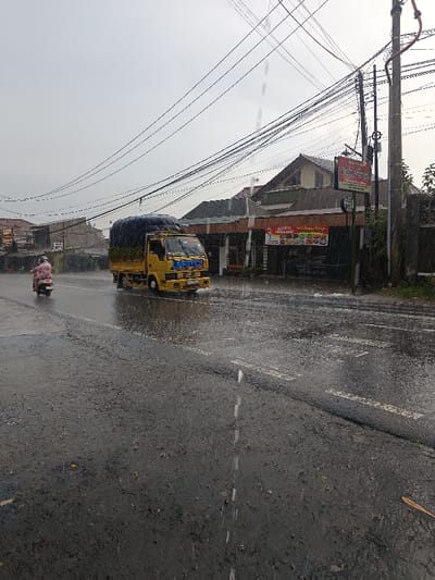 The scene depicts a street in Cugenang, Indonesia, during rainfall under an overcast sky. A wet asphalt road with visible white dashed lane markings extends from the foreground into the midground. A yellow medium-duty truck, bearing "LIVESTOCK" on its front bumper and carrying a dark, tarpaulin-covered load, moves right-to-left through the frame, generating tire spray on the wet surface. Behind the truck and slightly to its left, a person wearing light-colored rain gear rides a light-colored scooter in the same direction. On the right side of the street, commercial and residential buildings are visible, including one with a red sign advertising "Sate" and "Nasi Ayam Bakar Nasi Campur." A utility pole, heavily laden with numerous entangled electrical and communication cables, stands prominently on the right, further back. Additional structures and utility lines are present in the distant background on both sides of the thoroughfare.