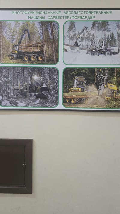 This media file is a photograph of a poster displayed on a wall, likely in a public space in Ukhta, Russia. The poster features four images of forestry equipment, specifically harvesters and forwarders. The text on the left side of the poster is in Russian and reads: "МНОГОФУНКЦИОНАЛЬНЫЕ ЛЕСОЗАГОТОВИТЕЛЬНЫЕ МАШИНЫ ХАРВЕСТЕР+ФОРВАРДЕР", which translates to "Multifunctional Logging Machines Harvester+Forwarder".
The top left image shows a white forestry machine with a large grapple arm extended, seemingly in a snowy forest. The top right image depicts a yellow forestry machine in action, cutting a tree with a chainsaw attachment, with sawdust flying. The bottom left image shows a yellow and black forwarder loaded with logs, moving through a forest. The bottom right image is a darker, more abstract view of a forestry machine in a forest, possibly covered in snow or debris, with headlights illuminated.
The overall scene is professional and informative, showcasing the capabilities of these specialized machines in a forest setting. The weather in the images appears to vary from snowy to perhaps wetter conditions. There are no people clearly visible as the main subjects, though one driver is discernible in the top right image. The poster itself is framed by a dark border. To the right of the poster, a portion of a wall and a dark, recessed panel are visible.