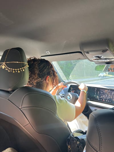 The image shows the interior of a car, likely taken from the backseat looking towards the driver. A woman with curly dark hair is seen from the side and back, gripping the steering wheel. She is wearing a light-colored t-shirt and a watch on her left wrist. A portion of a car's infotainment system is visible on the dashboard, displaying text that appears to be in French. A small black purse with a gold chain is placed on the passenger seat. The scene suggests the woman is driving during the daytime, as indicated by the sunlight filtering into the car and the bright exterior visible through the windshield and side mirror. The overall mood appears casual. The location context provided is Ougrée, Belgium.