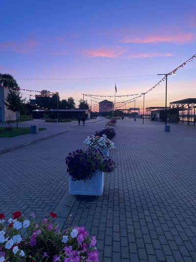 This image captures a sunset scene along a paved walkway in Sovetsk, Russia. The sky transitions from a deep blue at the top to shades of pink and orange near the horizon, with wisps of clouds scattered throughout. Along the walkway, strings of decorative flags and lights are strung between poles. Flower planters filled with purple, white, and red flowers line the left side of the path. In the distance, two figures walk away from the camera. The walkway is constructed of gray interlocking pavers. Structures, including buildings and covered seating areas, are visible on the right side of the path, and a body of water can be seen beyond them.