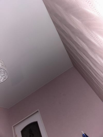 The image depicts an interior domestic space located in Charentsavan, Armenia. The scene is captured from an upward-angled perspective, showcasing a light-colored ceiling, walls, a portion of a door, a light fixture, curtains, and an iron.
The ceiling, occupying the upper left and central portion of the image, is a smooth, reflective, off-white or very pale grey surface, possibly a stretch ceiling, showing subtle, soft reflections. The walls are covered in a textured wallpaper of a uniform pale purple or dusty pink hue, displaying a fine, linen-like weave pattern.
In the upper left, a segment of a white, decorative lampshade is visible, featuring intricate geometric or fractal cut-out patterns. To the right, approximately one-third of the image width, light-colored sheer curtains or fabric are present, exhibiting a distinct wavy or rippled texture with subtle horizontal stripe patterns.
In the lower center-left, a white door frame encloses a dark-paneled door. The door's panel features a decorative pattern, possibly etched glass or an iron grille design. In the lower right corner, the front tip and a small section of a blue and white electric iron are visible, positioned against the pale purple wall.
No individuals are present in the scene, and no overt actions or interactions are occurring. The environment appears clean and well-maintained, illuminated by an unseen light source that casts soft, even lighting across the surfaces.