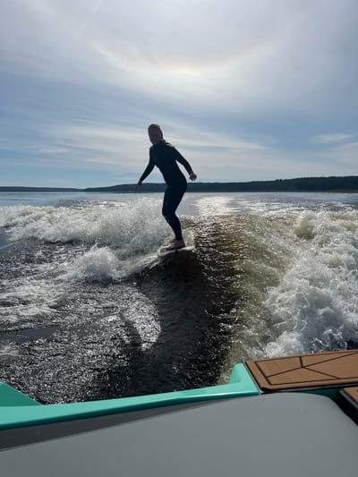 A person wearing a black wetsuit surfs on a wave generated by a boat. The person is positioned on the right side of the frame, riding a white surfboard with the nose slightly angled upwards. They are looking towards the left and have their arms outstretched for balance. The water is turbulent, with white foam and spray around the surfboard and extending behind it in a wake. The background shows a wide body of water, with a distant, tree-lined shore under a partly cloudy sky. A faint halo effect is visible in the sky. In the foreground, the back of a boat is visible, with turquoise and grey accents and a wooden deck section. The scene is set on the picturesque lake waters of Gromovo, Russia, a popular recreational area known for its natural beauty and outdoor activities.