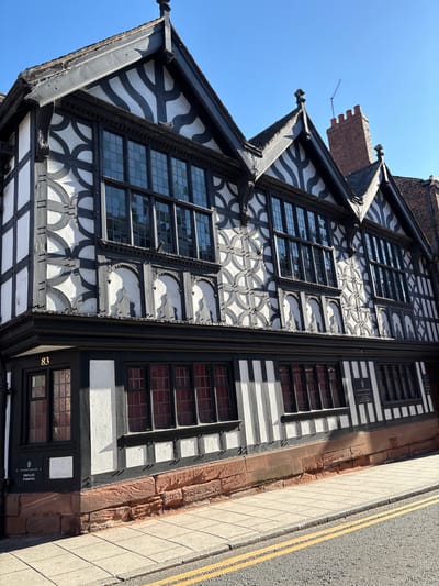The image captures Stanley Palace in Chester, United Kingdom, a notable example of Tudor architecture. The three-story building features black and white timber framing, a hallmark of the period. The framing is adorned with decorative patterns. Multiple paned windows are present across all floors. A brick chimney rises from the roof. The side of the building abuts a sidewalk and a street with double yellow lines. A sign near the base of the building reads "Stanley Palace Private Parking" along with the building's number, 83. The sky is clear and blue. The image is rotated slightly, with the top of the building leaning towards the left.