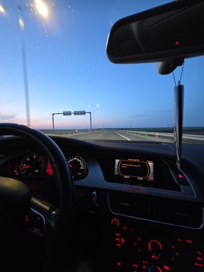 The image shows the interior of a car driving on a highway at dusk. The dashboard is visible, displaying information like speed (partially shown), engine RPM, and audio settings. The central console features illuminated buttons for radio, media, and climate control. The driver is in the car. Outside, the highway stretches into the distance with overhead signs and light poles. The sky is a gradient of colors indicating either sunrise or sunset.
It is impossible to tell if the image was taken in or near Dimovo, Bulgaria.