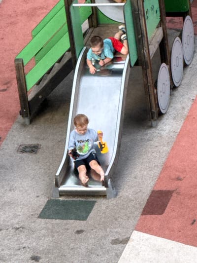 The image shows a playground slide with two young children on it. The child on the left is wearing red shorts and a blue shirt, with a toy in their hand. The child on the right is wearing a grey sweatshirt with the word "TOKYO" and a graphic on it, and black shorts. They are both at the bottom of the slide. The slide itself is made of metal and is part of a larger playground structure that includes green steps and platforms. The ground around the playground appears to be a textured concrete surface, with some reddish-pink areas that might be painted concrete or asphalt. There are also some circular grey objects at the top of the structure, possibly decorative elements or parts of another play feature. The overall scene suggests a sunny day, as indicated by the shadows. The children appear to be engaged in playing on the slide, with no strong indication of specific emotions other than the typical enjoyment of playground activities. The location is identified as Сухарево, Russia.