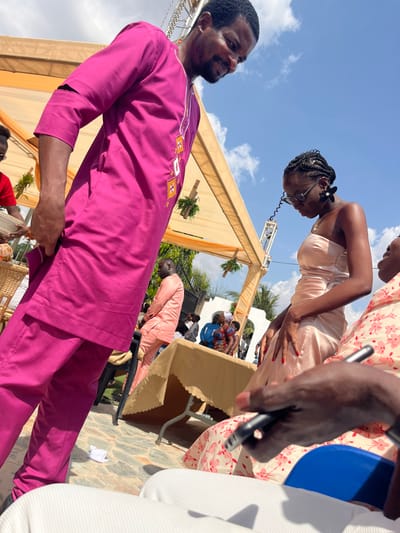 This outdoor image, captured in Kumasi, Ghana, depicts a social gathering under a beige fabric canopy on a sunny day. In the immediate foreground, a dark-skinned man with a beard, dressed in a bright magenta traditional Ghanaian tunic and matching trousers with white, yellow, and orange geometric embroidery, stands at an angle, looking downwards with a faint smile. His right hand is partially obscured, possibly tucked into his garment. To his right, a dark-skinned woman with braided hair and glasses, wearing a strapless light peach-colored satin-like gown, also looks downwards. Her fingernails are painted red, and her hands are clasped in front of her. In the lower right, the hands and lap of a seated individual are visible, holding a black smartphone, wearing a light-colored, floral-patterned top, and seated in a blue chair. A small woven basket containing light-colored spherical objects rests on a stand to the left of the main man. In the mid-ground, a dark-skinned man in a light peach traditional outfit is seated at a table covered with a scalloped beige tablecloth. The ground is paved with multi-toned stones, and a crumpled white paper lies near the main man's feet. The canopy structure features decorative orange fabric and hanging green planters. Several other guests are visible further in the background. The sky is clear blue with scattered white clouds.