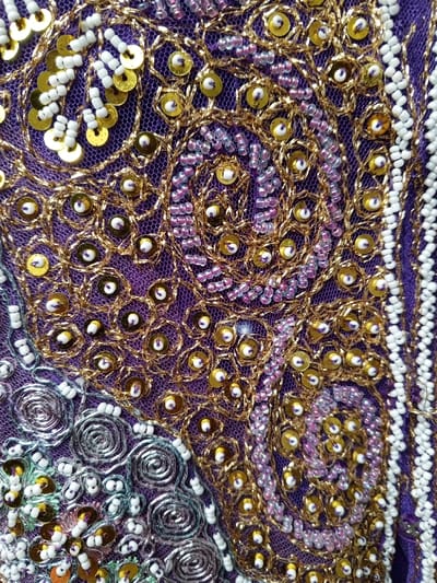 This close-up image displays a vibrant and intricately embroidered fabric, consistent with textile art found in places like Houaykhang, Laos. The primary subject is the detailed handiwork on the textile, featuring a rich interplay of colors and textures.
The base fabric is a deep, iridescent purple, possibly a net or mesh overlay on a solid material. This purple background is densely adorned with various embellishments. Gold-colored sequins are prominent, often secured by matching metallic gold threads that form swirling, organic, and paisley-like patterns across much of the surface. Within and around these gold designs, delicate strands of small, iridescent light purple or pink beads are meticulously stitched, adding another layer of texture and color.
Along the right side of the frame, and in smaller segments elsewhere, distinct patterns are created using opaque white beads, often arranged in neat lines or clusters. In the lower-left section, different decorative elements appear, including coiled silver metallic threads that form spiral motifs, interspersed with more white beads and gold sequins.
There are no people, visible activities, or events taking place in the image. The focus is purely on the craftsmanship of the embroidery. No specific time of day or weather conditions can be discerned, and there is no visible text. The overall impression is one of elaborate, detailed, and skillful textile artistry, showcasing traditional decorative techniques.