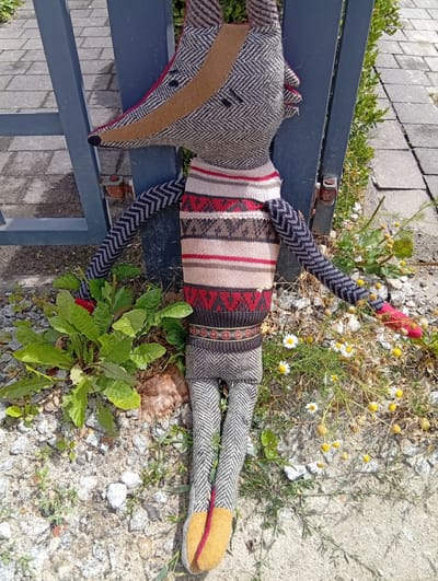 An overhead shot shows a stuffed toy resembling a fox or badger, standing upright against a dark blue metal gate. The toy is dressed in a patterned sweater with horizontal stripes in red, cream, and brown, and has a textured herringbone fabric for its head, arms, and legs. The toy's head is turned slightly to the left, revealing two black button eyes and a tan snout with a red trim. Its arms are outstretched, one resting on the gate, the other reaching towards a patch of green leafy plants and small white wildflowers. The ground beneath the toy is a mix of gravel, stones, and patches of concrete, with more wildflowers and dry grass scattered around. The scene appears to be outdoors, with the gate suggesting a boundary or entrance. The lighting indicates it is daytime, with sunlight casting shadows.