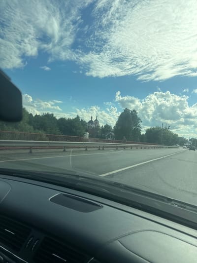 The image depicts a view from inside a vehicle, looking out onto a road with a bridge and a wooded area. The sky is bright blue with scattered white clouds, some of which are wispy and others are larger cumulus formations. The road appears to be a multi-lane highway, with traffic visible in the distance. A red railing runs along the side of the bridge, and beyond it, a dense line of trees separates the road from a small cluster of buildings, including a church with two domes. A lone figure is walking on the bridge. The interior of the car, including the dashboard and a portion of the windshield, is visible in the foreground. The location is identified as Kliukvennyi, Russia.
