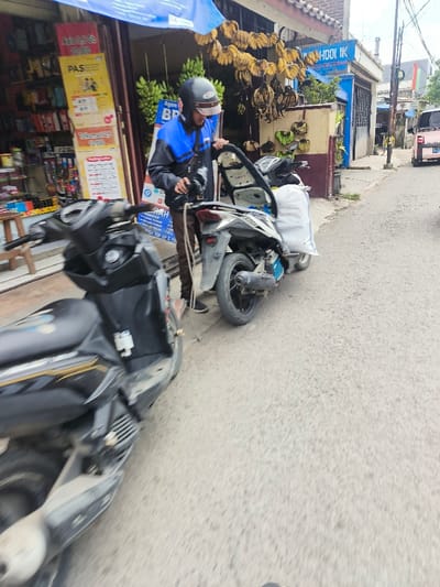 A person in a blue and black jacket and helmet is standing next to a parked white motorcycle with an open seat. A large white bag is secured on the back of the motorcycle. In the foreground, a blurred black motorcycle is visible. A building with bananas hanging outside is in the background, and a pink car is visible on the right side of the street.
