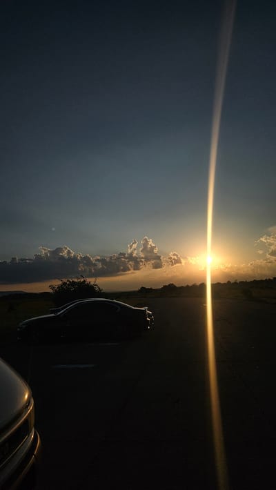 The image captures a sunset scene, likely located in the city of Buhovtsi, Bulgaria. The sun, a bright golden orb, is positioned in the middle-right portion of the frame, low on the horizon, emitting a pronounced vertical lens flare that traverses the entire height of the image. The sky above is a deep grey-blue, transitioning to warmer orange and yellow tones near the horizon. Scattered cumulus clouds are present across the mid-sky, exhibiting bright, illuminated edges from the backlighting of the setting sun, while their undersides remain in shadow.
An asphalt road occupies the foreground and mid-ground, appearing dark and featureless save for faint linear markings on its surface. Parked on the right side of this road is a dark-colored two-door coupe, heavily silhouetted against the bright horizon. The vehicle's make and specific features are obscured by shadow, though its profile and a small rear spoiler are discernible. In the bottom-left corner, the front bumper and grille of a second, lighter-colored vehicle, possibly an SUV or truck, are partially visible, also in deep silhouette.
The landscape beyond the road consists of undulating terrain with sparse, dark vegetation and bushes, all rendered in shadow. No people are visible within the frame. The scene is characterized by strong backlighting and high contrast, with deep shadows dominating the foreground and subjects, juxtaposed against the bright sun and illuminated clouds.
