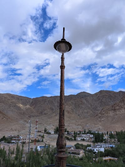 In the city of Phyang, India, this image captures a view of a village nestled at the base of a mountain range. In the foreground, a tall, ornate lamppost with intricate carvings stands as the primary subject. The lamppost is made of dark metal and features a decorative finial and a glass lamp housing.

Behind the lamppost, the landscape unfolds to reveal a collection of buildings, characteristic of a village or small town. The structures are a mix of white and earth-toned buildings, with some featuring red roofs. A prominent feature in the mid-ground is a cluster of two tall, modern communication towers, one white and orange, the other predominantly white. These towers stand out against the more traditional village architecture.

The village is surrounded by a verdant growth of trees, with a dense band of green foliage forming a natural border in the lower part of the image. In the background, rugged, barren mountains dominate the horizon. The slopes of these mountains are steep and covered in loose scree and rock, with a few faint tracks visible on their surfaces.

The sky above is a mix of bright blue patches and fluffy white clouds, suggesting a day with variable weather conditions. There are no people visible in the image. The overall scene depicts a juxtaposition of natural mountainous beauty, traditional village life, and modern infrastructure.