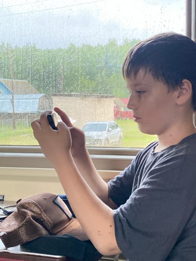 The media file shows a young person looking down at a phone, their hands holding the device. The person is wearing a dark grey t-shirt. To the left of the person is a window with raindrops on it, suggesting it is raining. Outside the window, blurred greenery and what appear to be buildings are visible. A brown baseball cap is draped over something behind the person. The lighting and rain on the window indicate it is likely daytime, possibly overcast due to the weather. The scene appears to be indoors, by a window, with a view of an outdoor environment. The person's focus on the phone suggests engagement with a digital activity. There is no visible text in the media file.