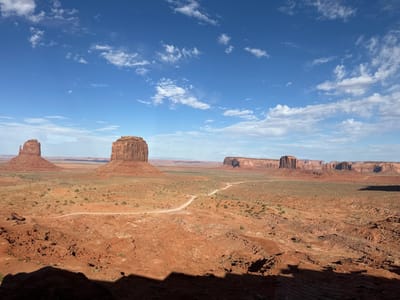 The image depicts a wide, arid landscape in Oljato-Monument Valley, United States, characterized by iconic sandstone formations known as buttes. The sky is a clear blue with scattered white clouds. The foreground is dominated by reddish-brown, rocky terrain, casting dark shadows. A dirt road winds through the middle ground, leading towards the buttes. In the distance, a series of majestic buttes stand tall against the horizon, with more of these geological formations stretching across the landscape. Sparse vegetation, consisting of low bushes and grasses, is scattered throughout the scene, indicating a desert environment. The overall scene conveys a sense of vastness and natural grandeur.