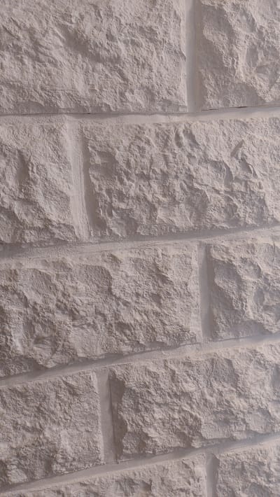 The image shows a close-up, textured view of a light-colored, rough-hewn stone wall. The stones are rectangular and laid in a staggered pattern, with visible mortar lines between them. The surface of each stone is uneven, with variations in depth and texture, suggesting natural stone or a realistic imitation. The lighting casts subtle shadows, highlighting the three-dimensional quality of the stone work. The background is entirely composed of this stone wall, and there are no people, animals, or other objects visible. The scene is static and appears to be an architectural detail. There is no information available to pinpoint its specific location within the city of Bad Laasphe, Germany.