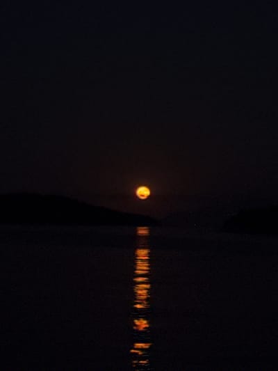 The image shows a dark scene with a luminous, orange circle, likely the sun or moon, low on the horizon in the left half of the frame. To the right of this circle, a dark, vertical shape cuts across the image, possibly a silhouetted object or a structure. In the lower right portion of the frame, a reflection of warm, flickering lights is visible on a dark surface, suggesting water. The overall impression is a tranquil, possibly twilight or night scene, with the faint glow of the celestial body and the reflected lights creating a serene atmosphere. The dark surroundings suggest a natural environment, potentially near a body of water. There is no visible text in the image. The Municipal Unit of Tolofona, Greece, provides a geographical context, hinting at a Mediterranean setting.