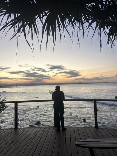 A landscape view shows a figure standing on a wooden platform, looking out at the water at dusk.
Environment: The scene is at dusk, with a yellowish-orange hue on the horizon where the sky meets the water. The water surface reflects the ambient light, with subtle wave patterns visible. The left side of the frame includes the silhouetted branches of a tree, while the foreground is the wooden platform with vertical supports.
People: A silhouetted figure, seemingly an adult, stands on the platform, facing the water. The person is wearing a striped shirt.
Objects: A surfboard leans against the structure on the right side of the frame.
Actions: The person is standing still.
Based on the landscape and the prevalence of beachfront structures, this image is likely taken in Coolangatta, Australia, due to its coastal location and the likely presence of surfing culture.