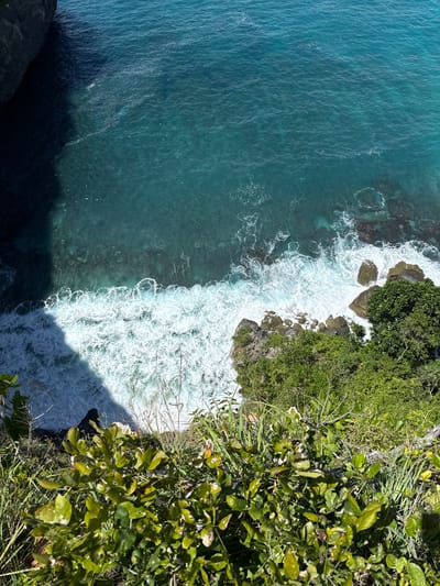 The image shows an aerial view of a coastline in Pejukutan, Indonesia. The turquoise ocean water occupies the left side of the frame, meeting the rocky shore. White waves crash against the rocks and the cliffside, creating a foamy surf. Lush green vegetation covers the cliffs, extending into the right side of the image. A shadow is cast by the cliff.