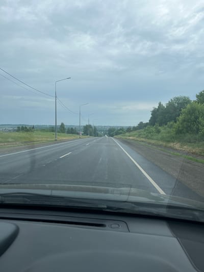 The image shows a view from inside a vehicle looking out at a two-lane asphalt road extending into the distance under a cloudy sky. The road has white lane markings and is wet, suggesting recent rain. Several streetlights are visible along the side of the road, accompanied by power lines. The landscape on either side of the road consists of grassy fields, trees, and some scattered buildings in the distance, indicating a rural or semi-rural setting. There are no visible people in the image. The front dashboard of the vehicle is partially visible in the foreground.