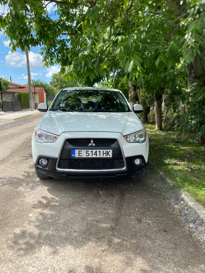 A white Mitsubishi ASX SUV is parked on a dirt road in Zagore, Bulgaria. The car faces forward. The front license plate reads "E 5141 HK" and has the Bulgarian "BG" emblem. The car is partially obscured by tree branches and leaves. A house with a red tiled roof is visible in the background. The sky is visible through the foliage and is blue with white clouds. The road is unpaved. A concrete curb runs along the right side of the road.
