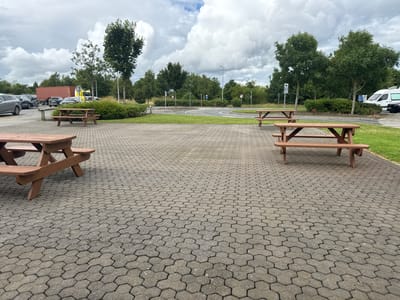 This media file shows an outdoor scene at Commons Cross, Ireland, featuring a paved area with hexagonal paving stones and several wooden picnic tables. In the background, there are trees, bushes, and a road with some cars and a large truck. The sky is cloudy, suggesting overcast weather. The time of day appears to be daytime, given the lighting.

The picnic tables are made of brown wood and are positioned on the paved area. One table is in the foreground, close to the camera, and another is visible further back. The surrounding area includes a grassy verge and more trees. In the middle ground, there's a road with a lane curving to the right. Several vehicles are visible, including cars and a large truck with a red shipping container. There are also some traffic signs and streetlights. The overall impression is of a rest stop or a park area adjacent to a road. No people are visible in the image.

Notable details include the hexagonal paving stones, the style of the picnic tables, and the type of vehicles, including the large truck. The overcast sky suggests a typical day in Ireland. There is no visible text of significance in the image.