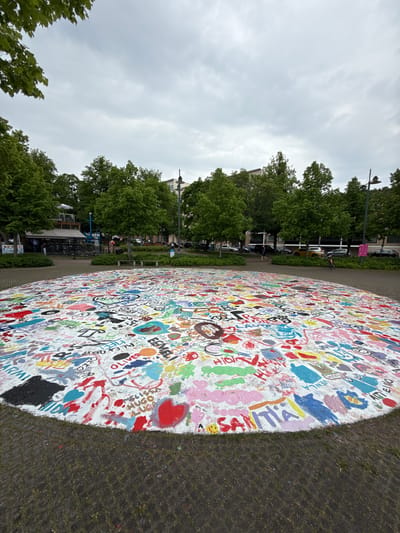 This image depicts a public space in Lappeenranta, Finland, under an overcast sky. The central feature is a large, circular, slightly raised white surface covered densely with multi-colored graffiti, including names, abstract shapes, hearts, and various scribbles. Visible names on the surface include "BUBBA" enclosed in a heart, "OLIVER," "ALEX," "EEMELI," "WILLIAM," "MELISSA," "HUGO," "SANNA," "IDA," "VENLA," "ATTI EINO," "OKKO," and "SAINTIA." A prominent black painted area is present on the lower-left edge of the white surface.
The ground immediately surrounding this painted surface consists of dark gray interlocking pavers with sparse green grass growing in the crevices. Beyond the paved area, numerous mature deciduous trees with vibrant green foliage form a natural barrier, obscuring the middle ground.
In the background to the left, through the trees, a multi-story building with light-colored walls is visible, featuring an outdoor seating area with white umbrellas and an awning, suggesting a café or restaurant. Indistinct human figures are present near this seating area. Further to the right, a dark lamppost with a curved arm stands tall, and behind the trees, additional multi-story buildings with light facades are visible. A street or parking area runs behind the trees on the right, populated by several parked cars of various colors (e.g., silver, yellow, dark). Two individuals are visible on a paved path in the middle ground on the right: one person in dark clothing walks from left to right, and another person in lighter clothing is further back near a bicycle. The sky is uniformly light gray, indicative of heavy cloud cover. Green leaves from an overhead tree branch are visible in the upper left corner of the frame.