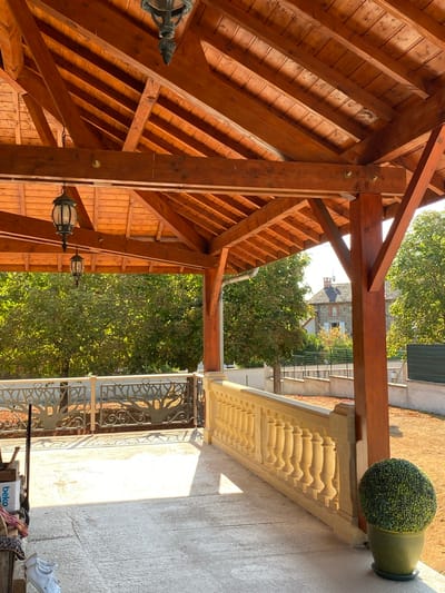 The image shows a covered outdoor patio area with a wooden roof structure. Two black lanterns hang from the wooden beams of the roof. A decorative metal railing with a tree motif runs along the edge of the patio, behind which is a stone balustrade. In the background, there are trees and a house with a tiled roof, suggesting a residential area in Courpière, France. Sunlight casts shadows across the concrete floor of the patio. A green potted plant is visible on the right side of the image. On the left side of the patio, a white pair of sneakers and some boxes are present.