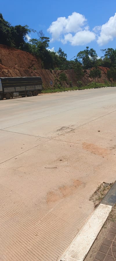 A large, tarp-covered truck is parked on the side of a paved road in Xayden, Laos. The truck is silver with a black tarp. To the left of the truck is a red dirt embankment covered in green foliage. The road appears to be a highway with a clear blue sky and white clouds overhead. The road is wide and appears to be in good condition. There are some small trees planted along the embankment to the right of the truck. The ground in front of the embankment has a rough, textured surface with some dirt patches.