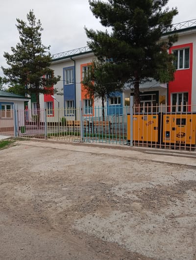 The image shows a colorful, two-story building, possibly a school or community center, behind a multi-colored metal fence. The building's facade is painted in alternating red, blue, orange, and white sections with white-framed windows. Two tall evergreen trees stand in front of the building, partially obscuring the view. To the right of the entrance are two yellow power generators. The foreground is a gravel-covered area. The sky is overcast. The location is indicated as Yukariyuz, Uzbekistan.