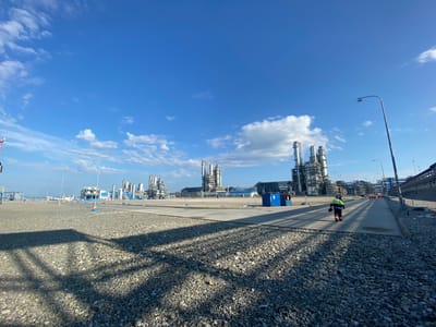An industrial landscape is depicted under a bright blue sky with scattered white clouds. In the foreground, a person in high-visibility workwear, including a yellow and blue jacket and orange pants, walks away from the camera on a paved pathway. The ground is covered in grey gravel. In the mid-ground, a row of industrial buildings with tall stacks and complex structures are visible. A blue utility building is situated near the pathway. Further in the background, more industrial facilities extend into the distance. The scene appears to be an outdoor industrial site. The image does not contain enough information to identify the specific city or country.