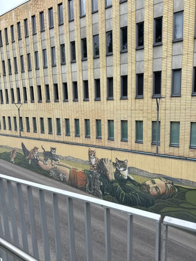 The image displays a multi-layered outdoor scene in Lappeenranta, Finland, featuring the "Man with Cats" mural by Jani Leinonen on a retaining wall along Valikatu street.
In the immediate foreground, a light grey metal railing with vertical balusters and a top handrail extends diagonally from the lower left to the lower right, partially obscuring the scene below.
Behind the railing, a large mural is painted on a light beige concrete retaining wall. The mural depicts a recumbent, bearded man with light hair and a fair complexion, wearing a dark green long-sleeved garment and reddish-brown lower garment. He is positioned on a green surface and looks directly upwards towards the viewer. Seven cats of various tabby patterns and colors surround him. One tabby cat rests on his chest, facing the viewer. A grey tabby cat is positioned on his raised left forearm, looking left. A larger tabby cat walks across his stomach/legs, moving left. A small tabby cat is on his lower right leg, walking left. An orange-and-white tabby cat is seated on his right hip, looking left. A grey-striped cat is near his left hand, looking right. Another small tabby cat is depicted further to the left on the painted green ground, walking left. The mural's background behind the man and cats consists of undulating green terrain below a lighter, muted yellow-brown upper section.
Above the mural and retaining wall, a multi-story building occupies the background. Its facade is primarily constructed of light yellow brick. The upper section of the building features rows of dark, horizontally oriented rectangular windows alternating with vertical bands of brick and ribbed grey panels. The lower section of the building, entirely brick, displays rows of vertically oriented rectangular windows with light-colored blinds visible internally. Two dark grey streetlights with single light fixtures are mounted on the building's facade; one is positioned on the upper left section, and another on the lower right section of the visible portion of the building.
A dark grey asphalt surface, consistent with a road or pedestrian path, is visible between the foreground railing and the mural wall. The scene is illuminated by soft, diffused light, indicating an overcast sky.