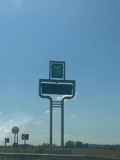 An eye-level, medium outdoor shot shows a large, stylized signpost with a graphic of an open book and a bird emblem under a clear blue sky with scattered clouds. The signpost is made of metal and has a modern design. To the left of the signpost are several traffic signs, including a circular sign with red and white markings and two square signs with red and white arrows indicating turns. In the background, there is a field and a line of trees, with power lines stretching across the horizon. The image is captured during daylight. The signpost is likely marking an entrance or a point of significance in the city of Krasnyi Oktyabr', Russia, as it features a local emblem and appears to be a landmark.