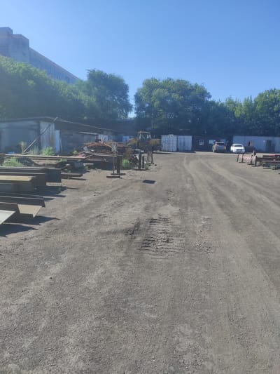 The image depicts an outdoor scene at a scrap metal yard, likely in Kyzyl, Kazakhstan. The ground is covered in gravel and dirt, with tire tracks visible in the foreground. Various stacks of metal beams and other scrap metal are scattered throughout the yard. In the background, there is a long, industrial-looking building with a light-colored facade and windows. Adjacent to the building, there are several shipping containers and a yellow construction vehicle, possibly a loader. Two cars are parked in front of the containers, one dark gray and one white. Trees line the background, and the sky is a clear, bright blue. No people are visible in the image.