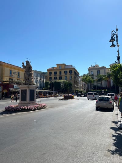 This image captures a sunny day in a piazza in Sorrento, Italy. In the foreground, a statue of a saint stands on a pedestal, surrounded by a bed of pink flowers. The statue depicts a figure with a halo, holding an object in its left hand and raising its right hand. Behind the statue, a street opens up with several cars and a few pedestrians. Buildings with multiple stories line the street, some featuring balconies and shops at ground level. Palm trees are visible scattered amongst the buildings. The sky is a clear, bright blue, indicative of fair weather. Several cars are parked or moving on the street, including a light-colored hatchback in the right foreground with a license plate that appears to read "FT 595CE". A lamppost with multiple lights stands on the right side of the image. People are seen walking on the sidewalks and near the statue, some appearing to be tourists. Outdoor seating for restaurants is visible. The overall scene suggests a lively public space in a European city.