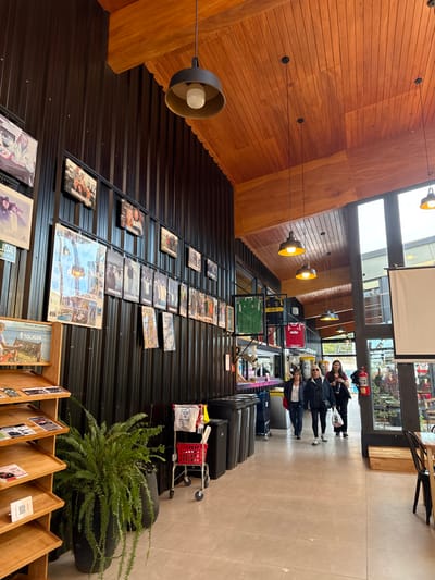 The image depicts the interior of a commercial or public building located in Tolhuin, Argentina, as indicated by a visible sign on a display shelf. The space features a high, angled ceiling constructed from light brown wooden planks and dark, vertically corrugated metal walls. The flooring consists of light-colored square tiles.
On the left side, a multi-tiered wooden display shelf holds brochures, one clearly displaying "TOLHUIN" and a landscape image. A large potted fern in a dark planter is situated in front of this shelf. The dark corrugated wall above is extensively covered with framed photographs and posters arranged in horizontal rows.
In the mid-ground, a red shopping cart or utility trolley with white bags attached is parked next to a series of dark-colored bins. Further along the wall, framed sports jerseys, including green and red ones, are displayed.
The right side of the scene features large windows extending towards the ceiling, allowing natural light and revealing an exterior view. A white projection screen is partially visible, hanging from the ceiling near these windows. A red fire extinguisher is mounted on a pillar closer to the windows.
Overhead, multiple black pendant lights with gold interiors and exposed bulbs hang from the wooden ceiling, providing illumination.
Three adult females are walking from the mid-ground towards the foreground on the right side of the tiled aisle. One woman, on the left of the group, wears a light top and dark pants. The middle woman wears a dark jacket, dark pants, and has sunglasses on her head, carrying a light-colored bag. The woman on the right wears a dark top and dark pants, also carrying a light-colored bag. Their action consists of walking through the interior space.