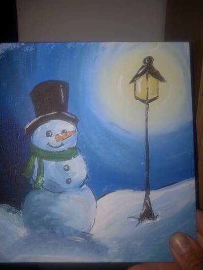 The image is a painting of a snowman standing in the snow at night. The snowman is wearing a black top hat and a green scarf, and it has a carrot for a nose and two black dots for eyes. A lamppost with a lit yellow lantern stands behind the snowman. The background is a dark blue sky with a bright yellow glow emanating from the lantern. The snow is depicted with white and light blue brushstrokes. The painting is held by a person's hand, partially visible at the bottom right. The setting appears to be a winter scene, likely in a park or garden. There is no visible indication that this scene is located in Kielce, Poland.