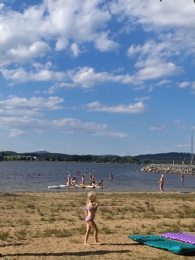 This is a landscape orientation, outdoor, daytime, portrait-oriented photograph. The scene depicts a body of water with a sandy shore. Several people are visible. On the shore, a young girl in a purple bikini lies on the sand. Further back, a group of people are on paddleboards in the water. One person is standing on a paddleboard, holding a paddle. The sky is blue with scattered white clouds. A rocky embankment is visible in the background, with a line of trees and hills beyond that. Two inflatable mats, one purple and one teal, are lying on the sand in the foreground. The lighting suggests it is a sunny day, likely late afternoon or early evening due to the long shadows. The overall atmosphere appears to be one of leisure and recreation. There is no visible text in the image.