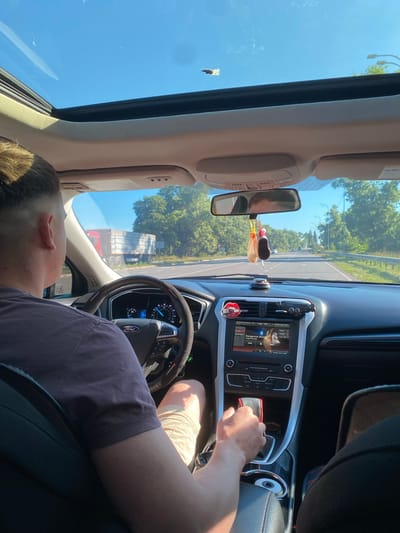 The image depicts the interior of a car, viewed from the passenger's perspective, as the driver, a person with short hair and a purple t-shirt, operates the vehicle on a sunny day. The driver's hands are on the steering wheel, and the car's dashboard and infotainment system are visible. The infotainment screen displays the song "Dangerous Woman" by Ariana Grande. A truck is seen in the distance on the road ahead, suggesting the car is on a highway or a major road. Trees line the sides of the road, indicating a rural or suburban setting. The car has a sunroof, which is open, allowing natural light to enter the cabin. The scene appears to be set in Herasymivka, Ukraine, based on the provided context.