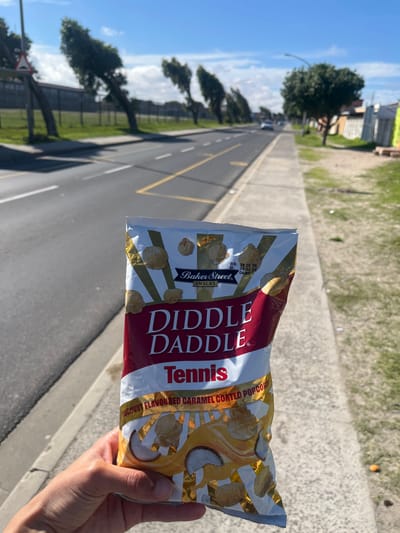 A hand holds a bag of Tennis Diddle Daddle Coconut Flavoured Caramel Coated Popcorn. In the background, an asphalt road stretches into the distance, marked with yellow lines. Trees line the left side of the road, and buildings are visible in the far distance. The sky is partly cloudy. The environment suggests an urban area, possibly located in Mitchells Plain, South Africa.