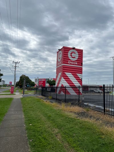 This image captures an outdoor scene under an overcast sky, likely in Hamilton, Australia. The dominant subject is a tall, vertical structure constructed from stacked red shipping containers. The containers feature large white diagonal stripes and are branded with a white circular "C" logo and the word "Dock" written vertically in white letters.

To the left of this container structure, behind a black metal fence, runs a multi-lane road or highway, devoid of visible traffic. The immediate foreground on the right shows a strip of green grass next to a concrete sidewalk that curves slightly to the left as it recedes into the distance. Patches of dry, brown grass are interspersed within the green.

In the mid-ground, beneath the level of the main road and behind the container structure, several smaller signs are visible. One prominent red sign also features the "Dock" branding, and another sign reads "VIBES SHOPS DECK C" with the number "217" on an adjacent sign. These indicate a commercial or business precinct. A utility pole with multiple power lines is visible on the far left side of the foreground, extending upwards into the cloud-filled sky. The sky is predominantly grey with visible streaky cloud formations, suggesting a cloudy day. There are no people visible in the image.