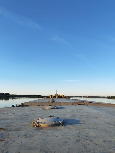 A view from the deck of a barge on a calm river under a clear blue sky. The barge's deck is concrete, weathered and worn, with several circular, metal hatches visible. In the distance, the barge stretches out, lined with yellow cylindrical objects, possibly part of its cargo or equipment, and a yellow crane. The river, reflecting the sky, is bordered by a lush green tree line on the far bank. A few small structures and a red buoy are visible on the distant shore. The sun is low in the sky, casting a warm light that illuminates the scene, suggesting it's either early morning or late evening. The overall mood is peaceful and still. The location context is Нифантово, Russia.