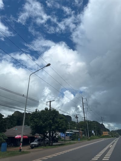 The image shows a rural roadside scene in Ban Thung, Thailand, under a dramatic sky with a mix of blue patches and heavy gray clouds. A paved road with white dashed lines runs diagonally from the bottom right towards the middle left of the frame. Along the left side of the road, there is a row of utility poles supporting numerous overhead wires. A tall street lamp stands prominently on the left. In the mid-ground, there is a building with a dark, sloped roof, partially obscured by trees. A pickup truck is parked in front of the building. Further down the road, more utility poles and trees line the landscape, leading to a mountainous backdrop. The overall atmosphere is overcast, suggesting potential rain.