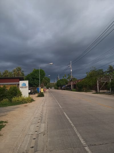 The image depicts a street scene in Moo 4, Thailand, characterized by overcast skies and a sense of impending rain. The road, made of gray asphalt with a double yellow center line and a broken white line marking the edge of the lane, stretches into the distance. On the left side of the road, a white sign with the words "DANU COCO Pool Villa KOH PHANGAN" is visible. Vegetation is abundant on both sides of the road, with trees and bushes lining the sidewalks and yards. Power lines crisscross above the street, supported by utility poles. The environment suggests a rural or semi-rural area. The image is devoid of people, but there are motorcycles parked behind the sign.