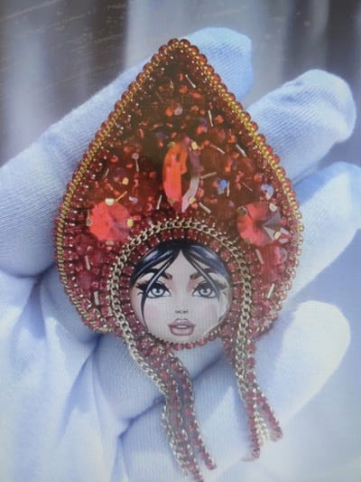 A detailed, embroidered brooch depicting a woman's face wearing a red, jeweled headdress is held by a gloved hand against a light blue fabric background. The brooch is teardrop-shaped and adorned with numerous red beads and rhinestones, with gold chain accents. The face on the brooch has dark hair, blue eyes, and red lips. The image is taken from a slightly overhead perspective. The location in Pospelikha, Russia is not discernible from the image itself.