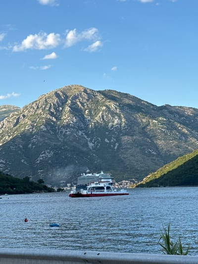 A ferry boat with white upper decks and a red hull navigates a body of water in front of a large, green, and rocky mountain. The sky is blue with scattered white clouds, and a small bird is visible flying in the upper left quadrant. The water has ripples, and several buoys are floating on its surface. A small village is nestled at the base of the mountain on the right side of the frame, with some vegetation visible in the foreground on the right. The scene appears to be in Jošice, Montenegro.