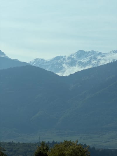 The image shows a mountain range, likely in Bulgaria, based on the implied location in Balanovo. The left side of the image features a snow-capped mountain with visible rock formations. The right side reveals a densely forested mountainside, possibly indicating lower elevations. The sky appears light, suggesting a clear or slightly hazy day.